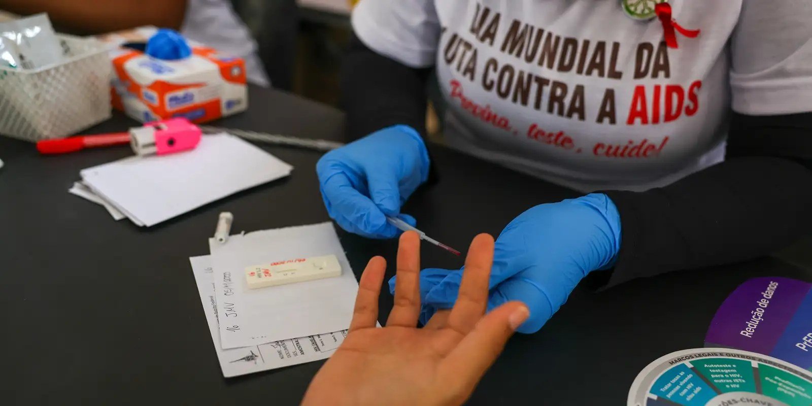 Profissional de saúde com luvas faz teste rápido de HIV no dedo de uma pessoa durante ação de prevenção