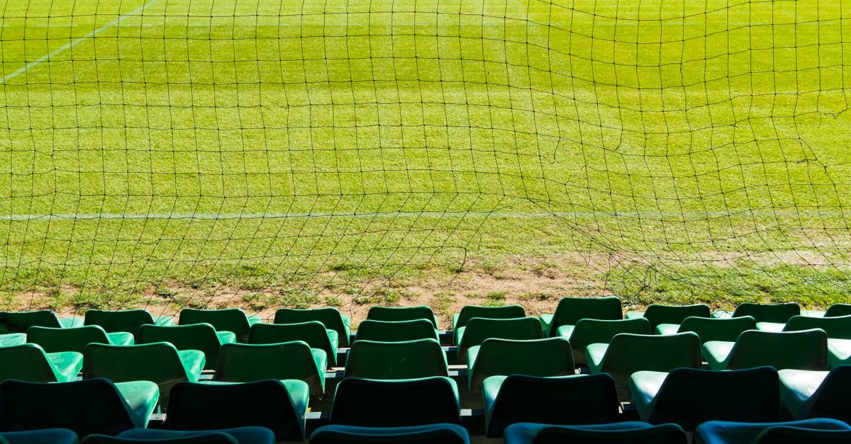 Gramado de estádio visto através da rede, com arquibancada verde vazia em primeiro plano