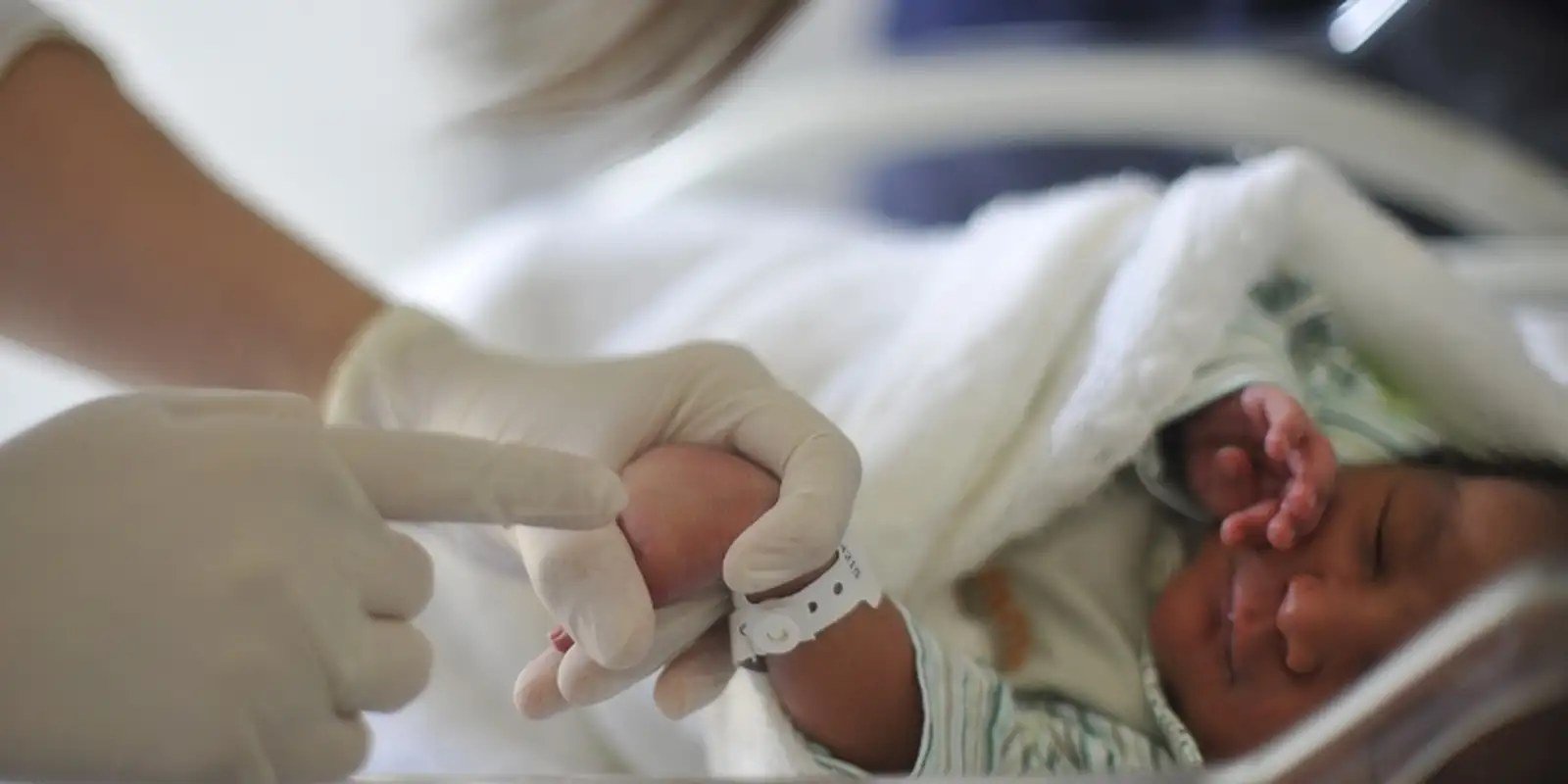 Recem-nascido enrolado em manta no berco de maternidade, com profissional de saude ao lado