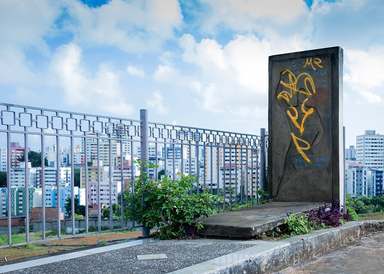 Lápide de Carlos Marighella, em concreto e com pichações, num cemitério de Salvador, com prédios da cidade ao fundo