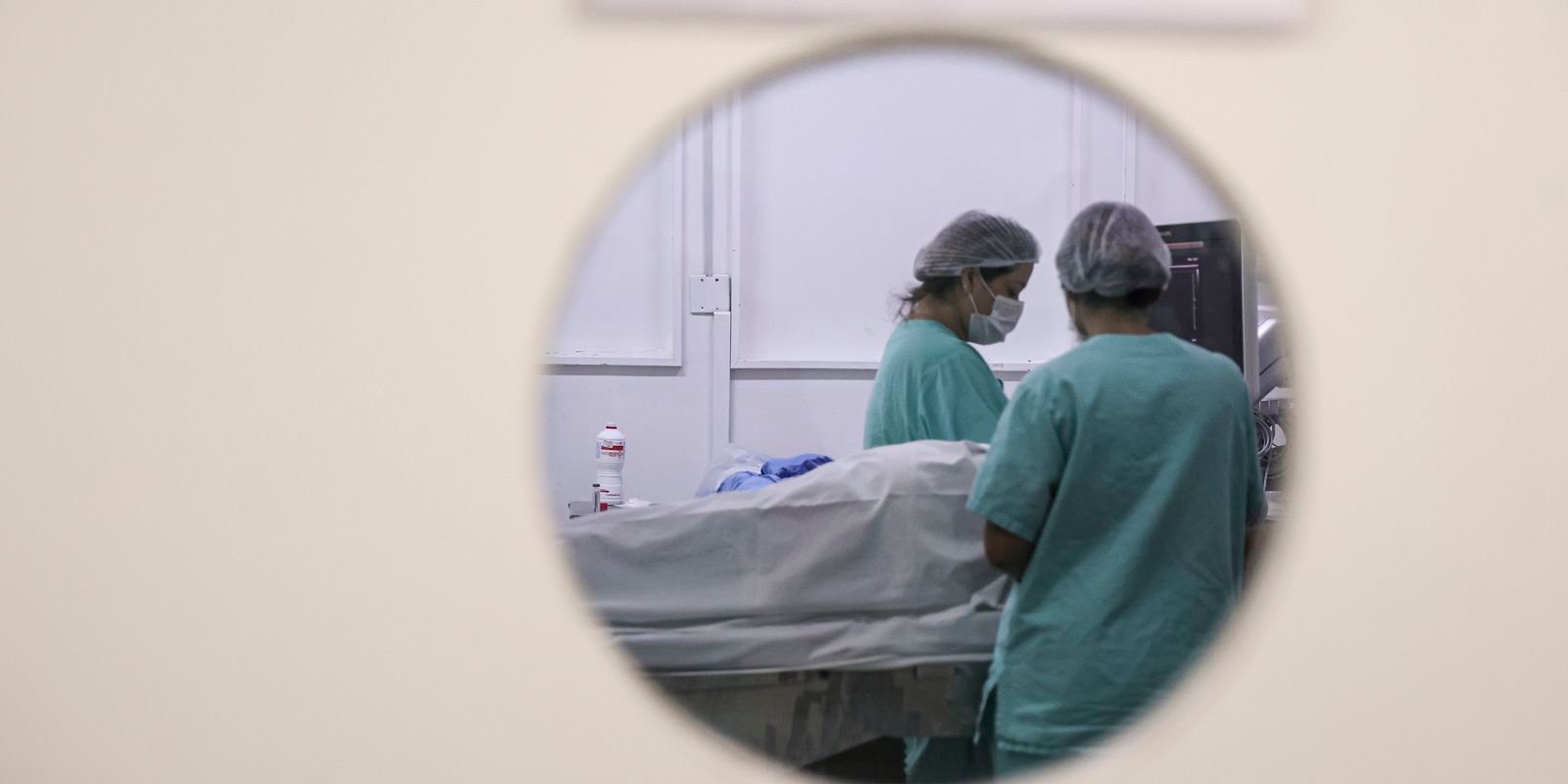 Profissionais de saúde atendem paciente em centro cirúrgico, vistos por abertura circular da porta