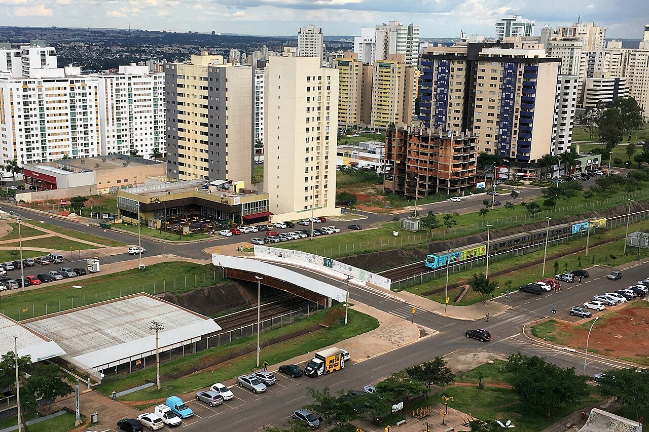 Trem do Metrô-DF em trecho a céu aberto, visto do alto, com prédios de Brasília ao fundo