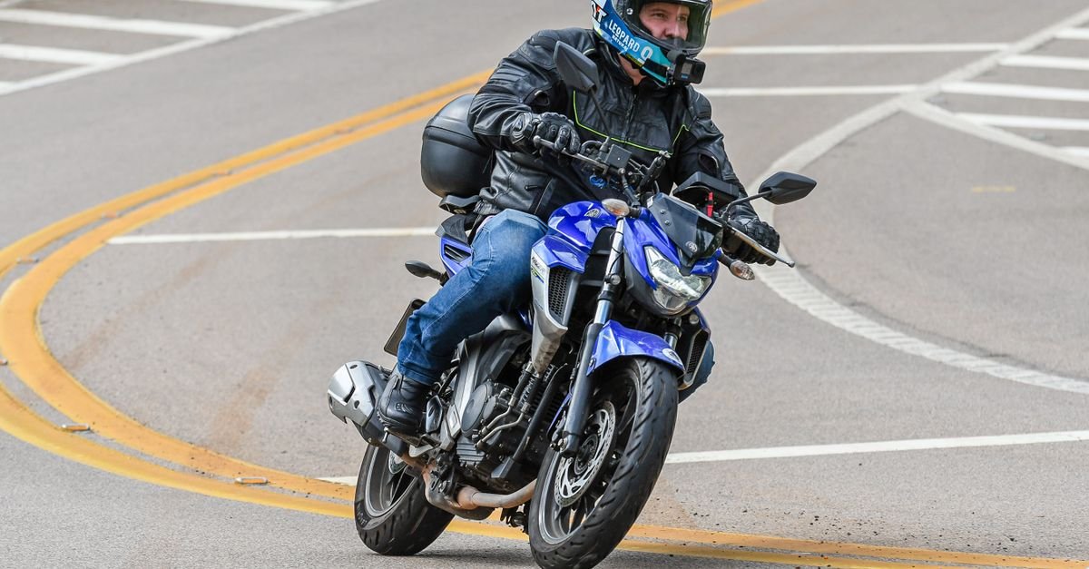 Motociclista com capacete e jaqueta de proteção faz curva em via asfaltada com faixas de sinalização amarela e branca