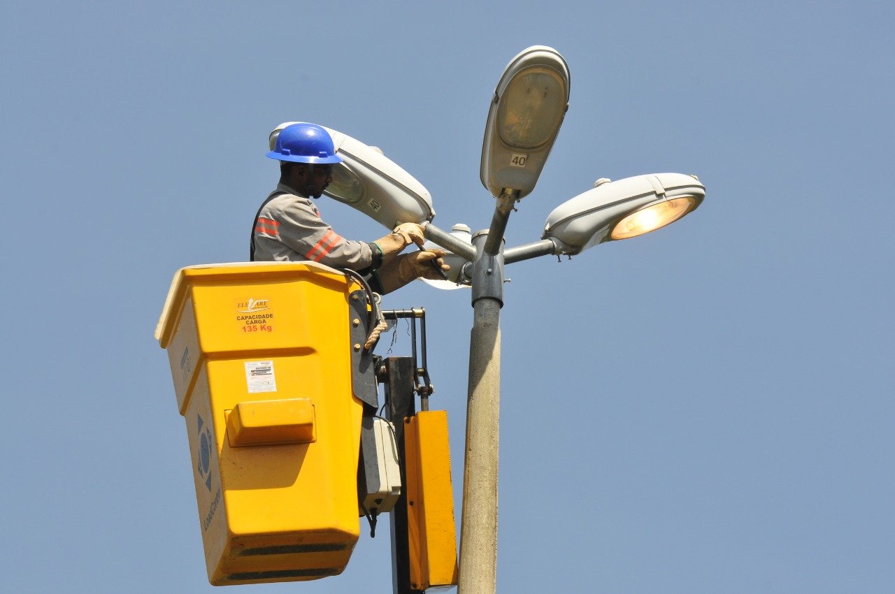 Eletricista de capacete azul faz manutenção em luminárias de poste sobre cesto elevatório