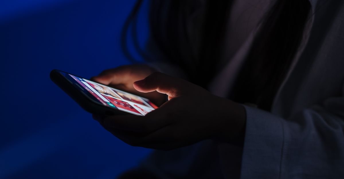 Mãos segurando um celular com a tela acesa em um ambiente escuro, sem rosto identificável