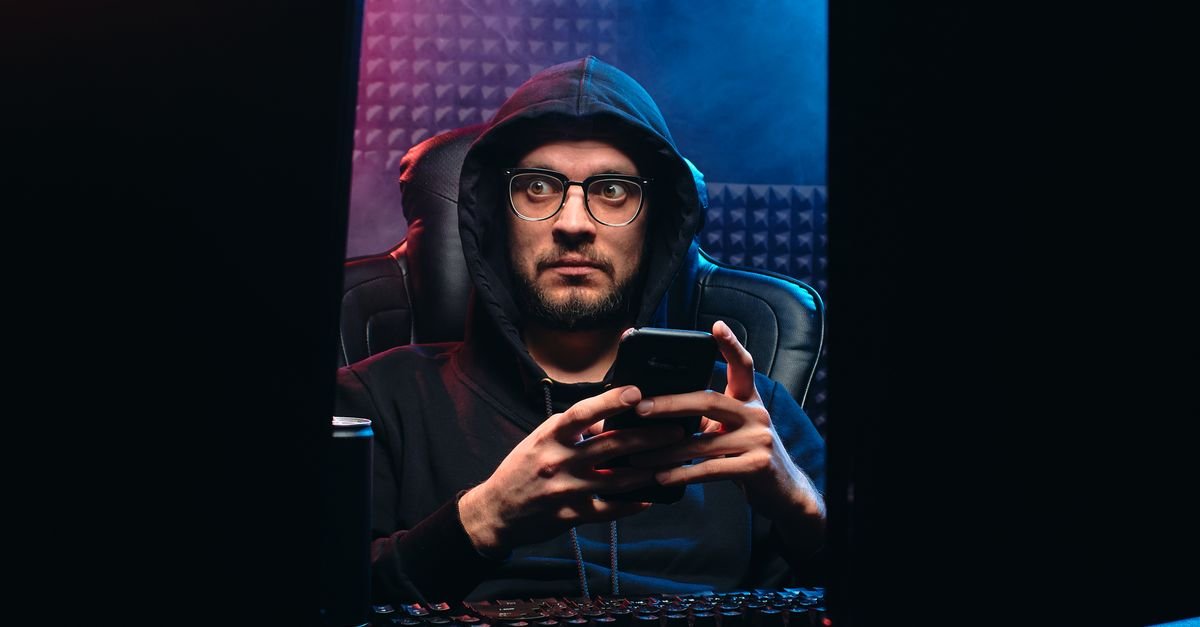 Homem de capuz mexe em celular em ambiente escuro, em imagem que ilustra crime virtual