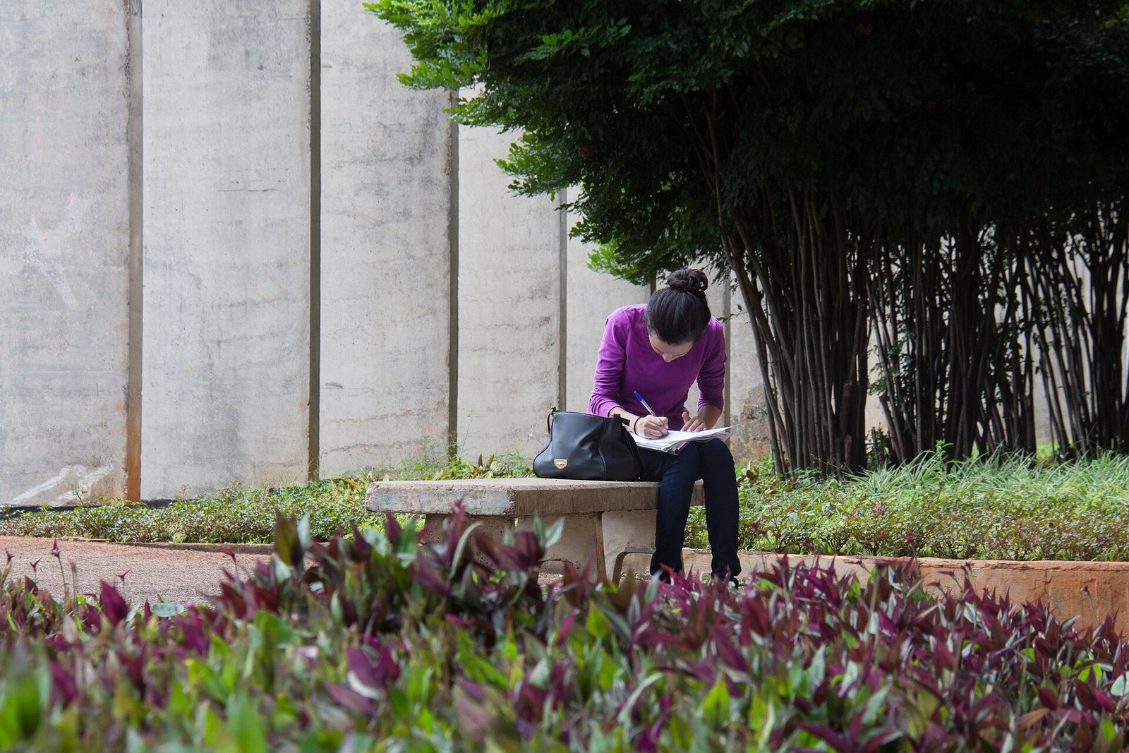 Estudante sentada em banco de concreto no campus da Universidade de Brasília escrevendo em um caderno apoiado no colo