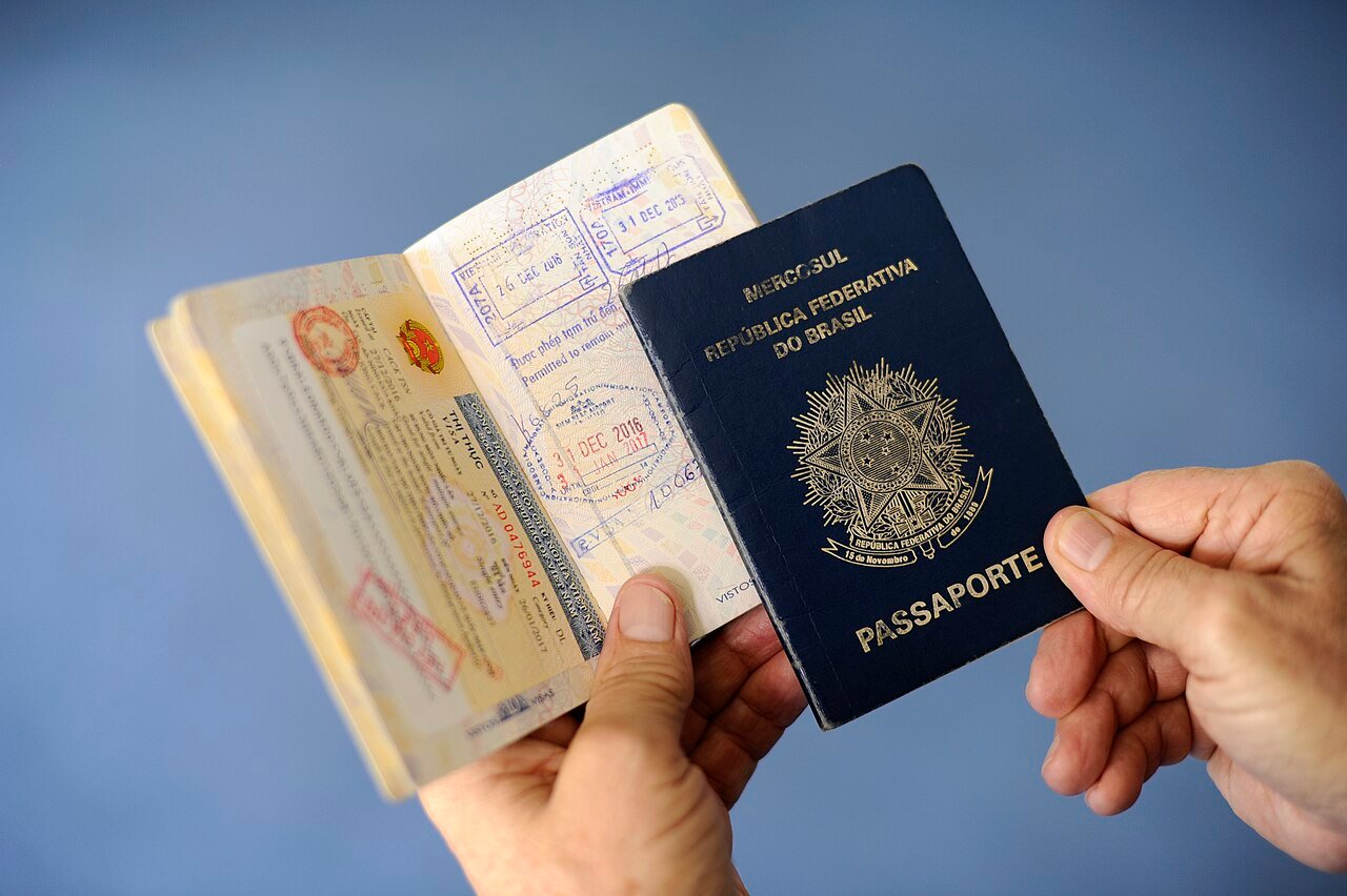 Mãos seguram um passaporte brasileiro fechado ao lado de outro passaporte aberto com carimbos de vistos
