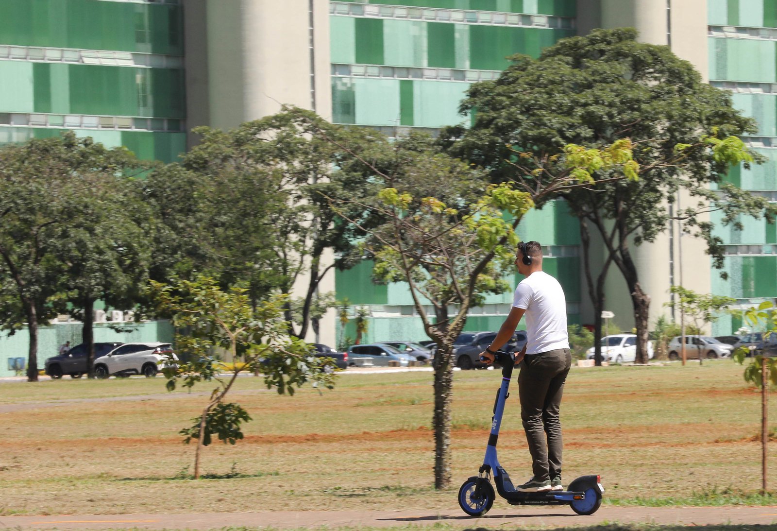 Homem anda de patinete elétrico azul da JET em gramado com prédio público ao fundo em Brasília
