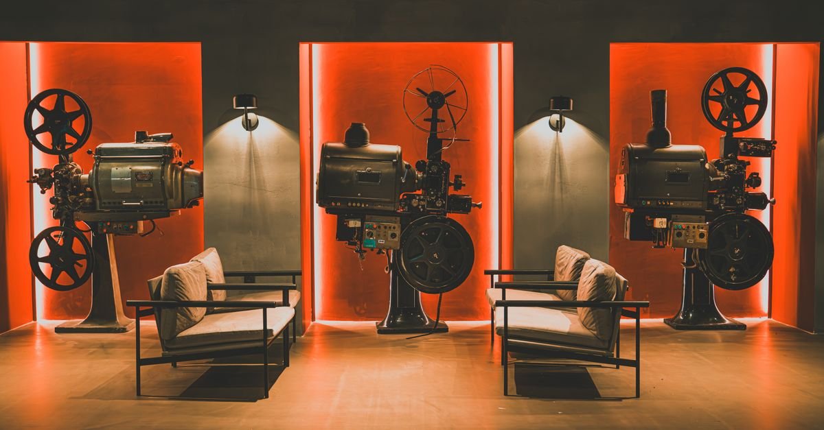 Projetores de cinema antigos expostos em uma sala com poltronas e iluminação alaranjada