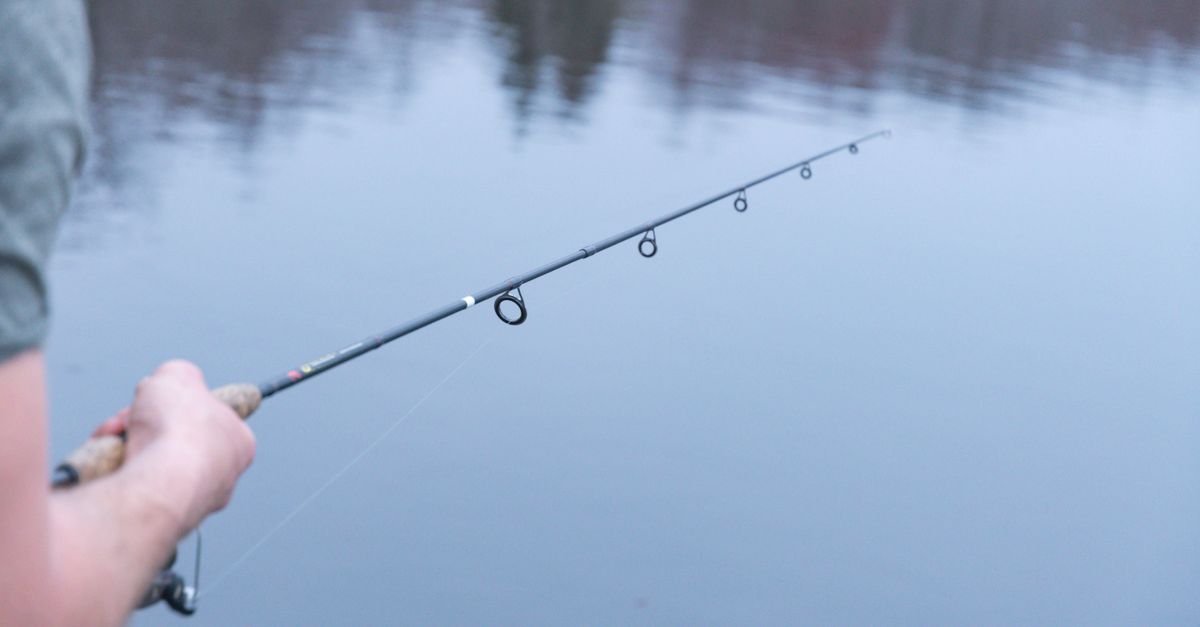 Mão segurando uma vara de pesca esportiva sobre a água parada de um lago