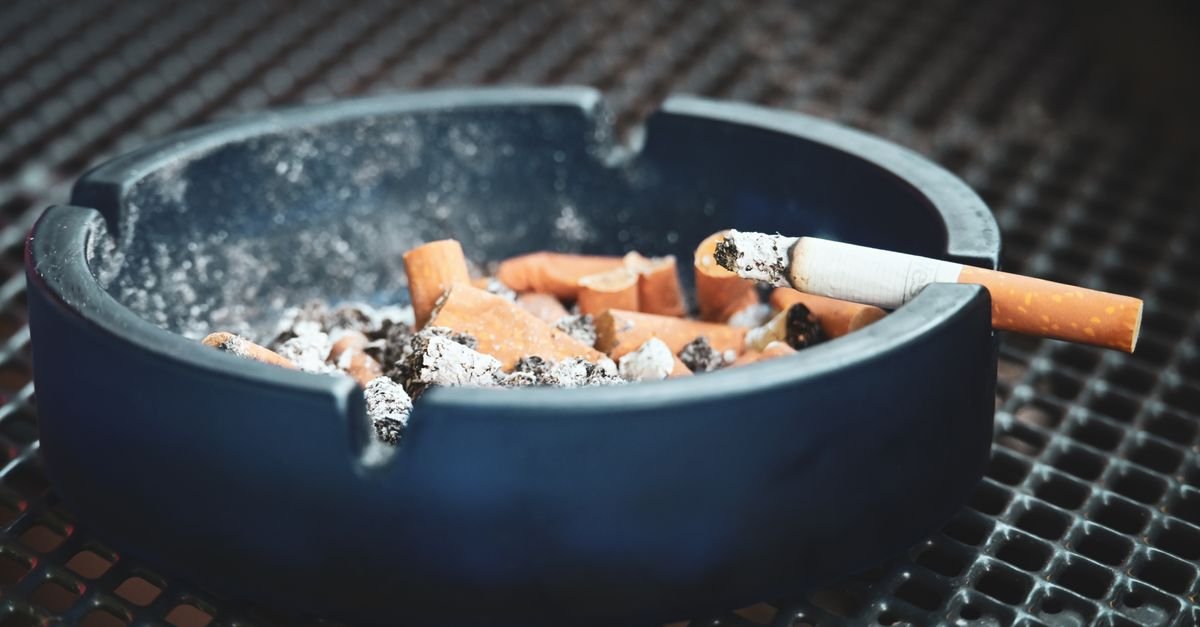 Cinzeiro azul cheio de pontas de cigarro e cinzas com um cigarro aceso na borda