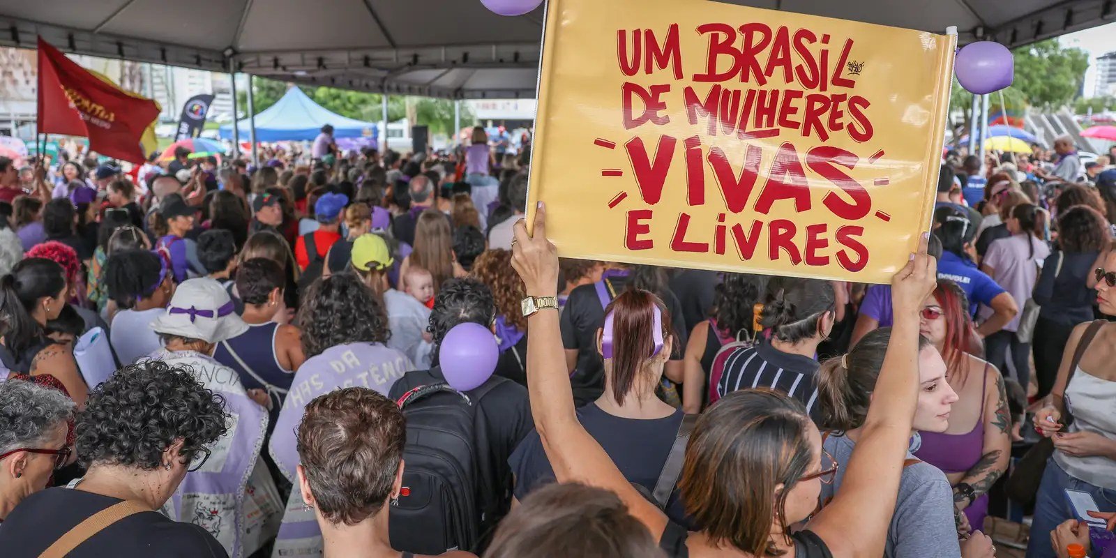 Mulheres reunidas em ato publico; uma delas ergue cartaz com a frase Um Brasil de mulheres vivas e livres