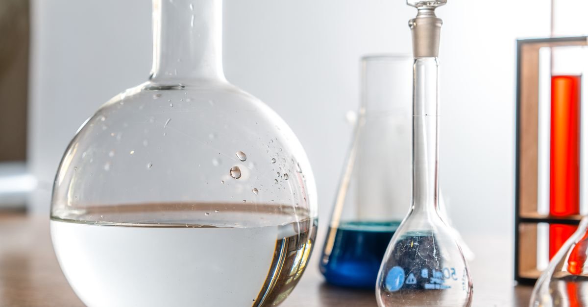 Vidrarias de laboratório sobre bancada, com balão volumétrico transparente em primeiro plano e frascos com líquidos coloridos ao fundo