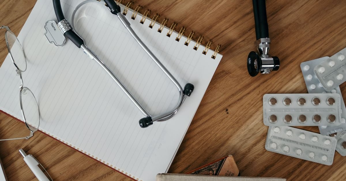 Estetoscópio, óculos e caderno sobre mesa de madeira ao lado de cartelas de comprimidos