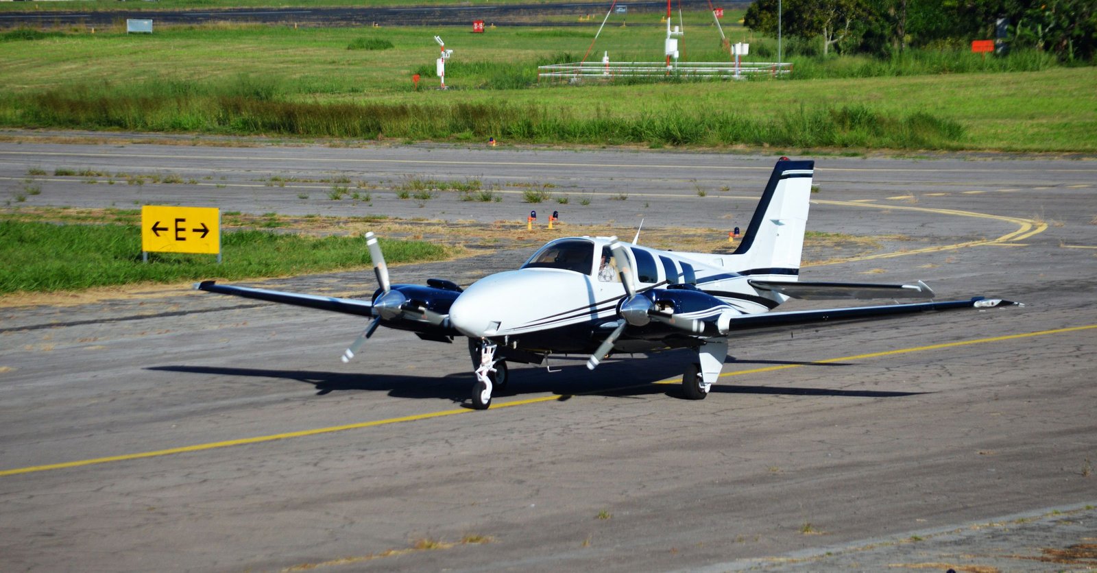 Aeronave bimotor de pequeno porte parada na pista de taxiamento de um aeródromo, com vegetação ao fundo