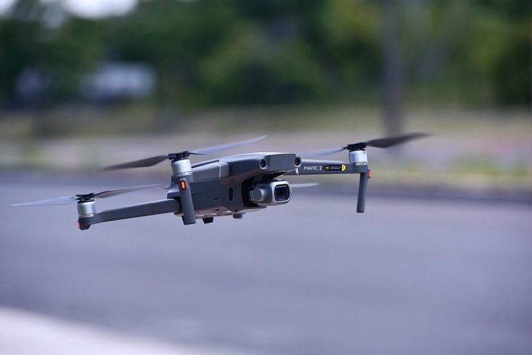 Drone de quatro hélices em voo baixo sobre uma via asfaltada, com vegetação desfocada ao fundo