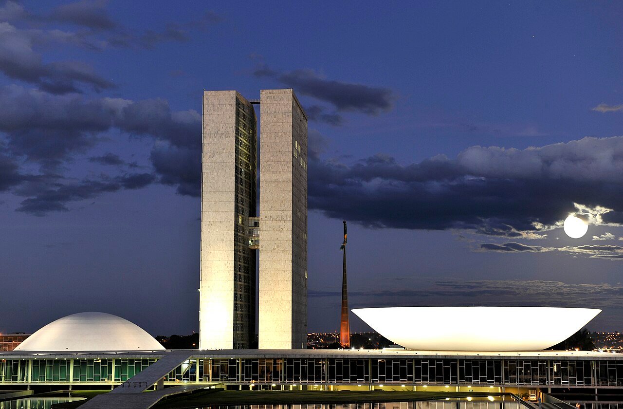 Congresso Nacional iluminado à noite, com as cúpulas do Senado e da Câmara e lua cheia ao fundo