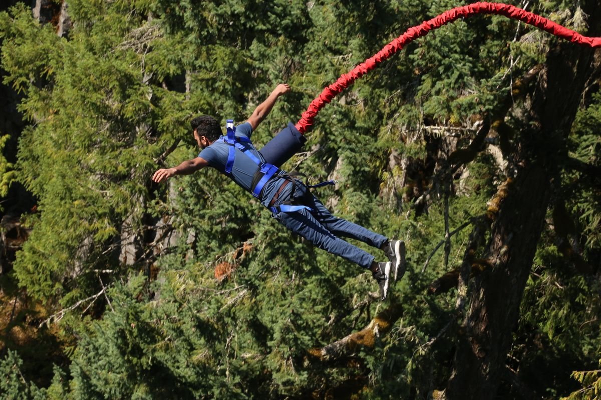 Homem salta de bungee jump preso a uma corda elástica vermelha sobre uma área de mata