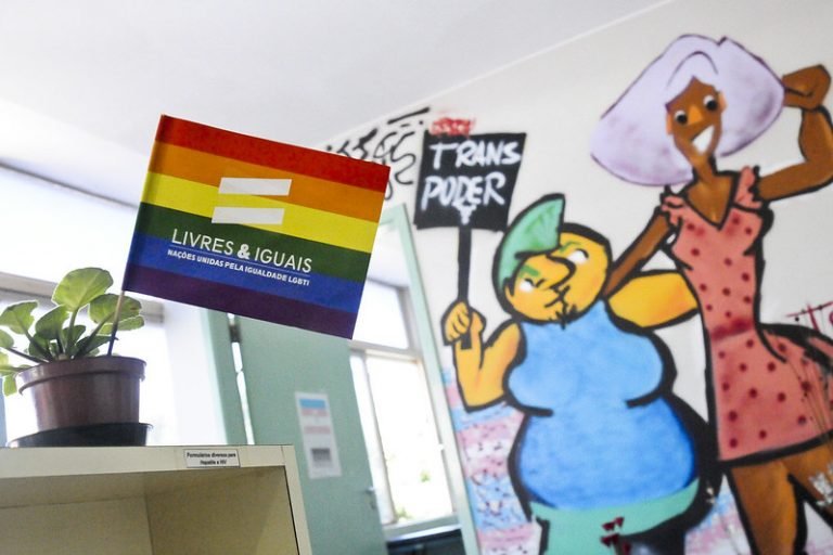 Bandeira de arco-íris com a inscrição Livres e Iguais sobre balcão de unidade de saúde, com mural colorido ao fundo