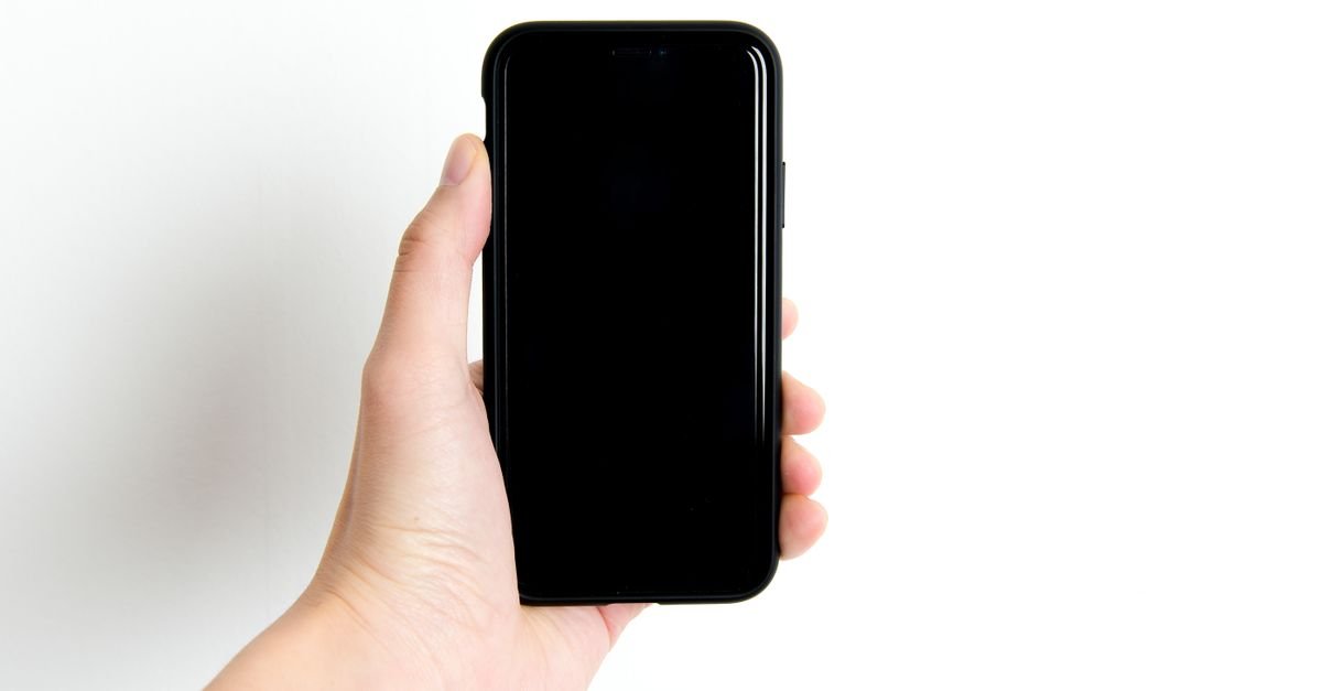 Mão segurando um smartphone preto com a tela apagada, sobre fundo branco