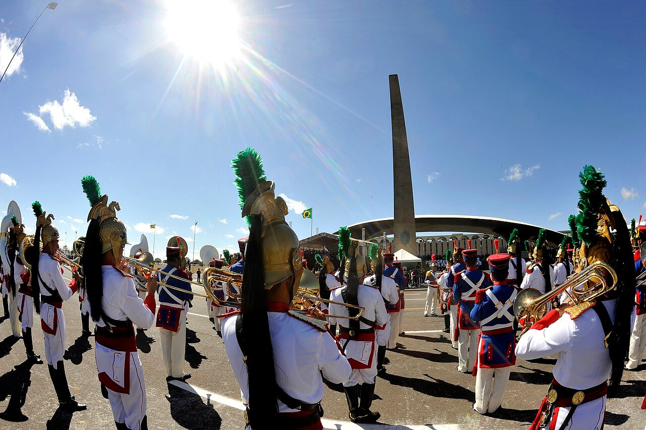 Banda militar em formação durante desfile no Quartel-General do Exército, em Brasília, com o monumento e o arco da praça ao fundo