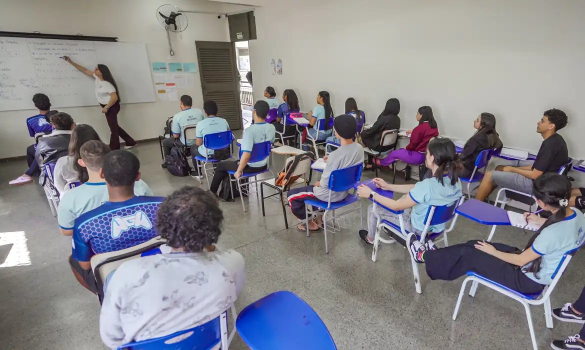 Sala de aula com estudantes sentados em carteiras e professora escrevendo no quadro branco