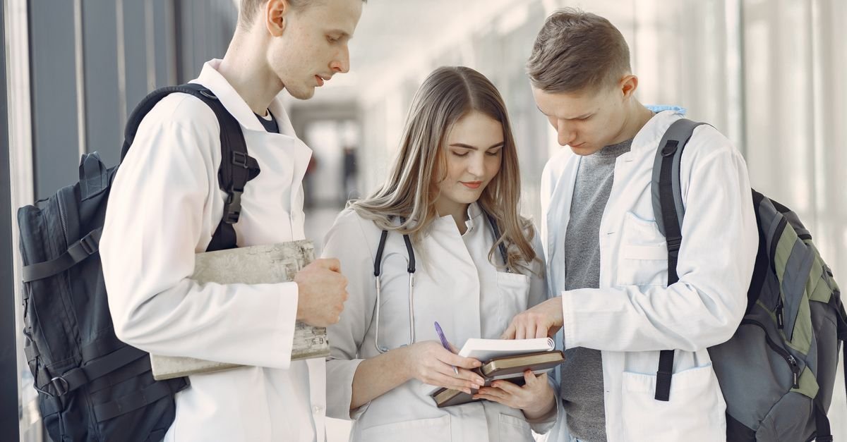 Três estudantes de medicina de jaleco branco conversam em corredor de hospital enquanto consultam um livro, imagem ilustrativa