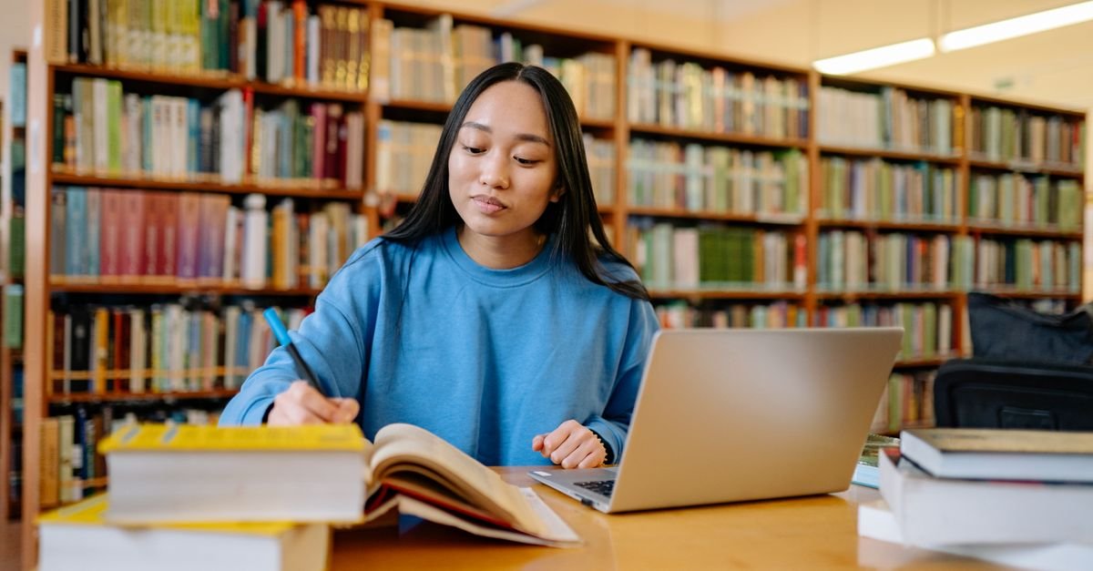 Estudante escreve em caderno ao lado de livros e notebook em biblioteca