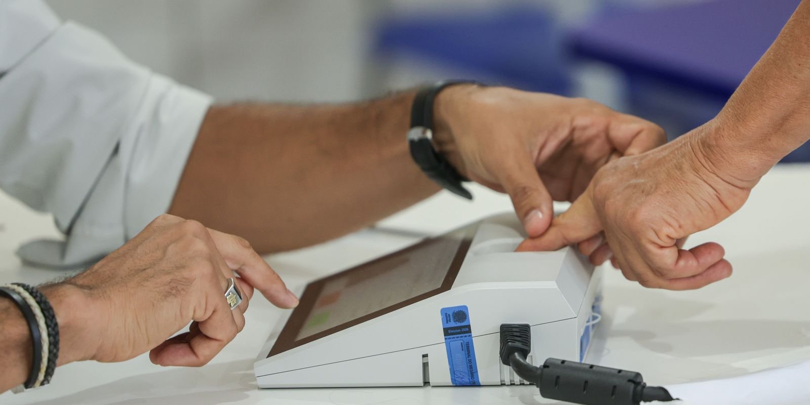 Mãos de um mesário e de um eleitor no terminal de identificação da seção eleitoral, com selo Eleições 2026