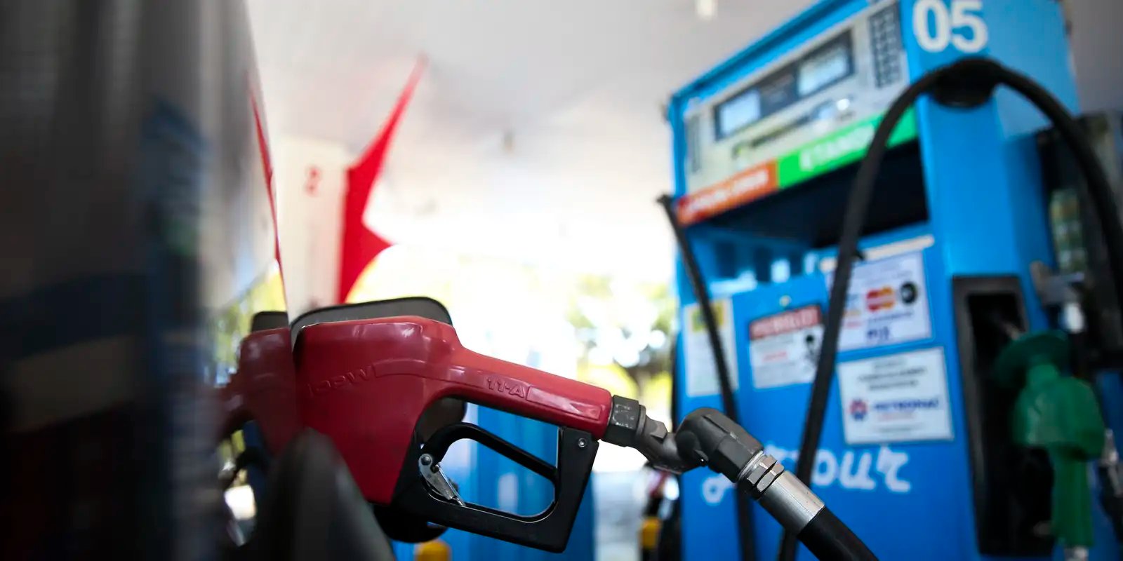 Bico de abastecimento vermelho encaixado no tanque de um carro, com bomba de combustível azul ao fundo em posto brasileiro
