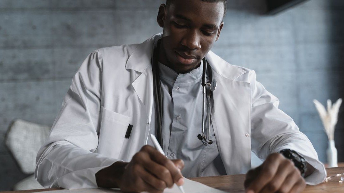 Médico de jaleco branco com estetoscópio escrevendo em uma mesa de consultório