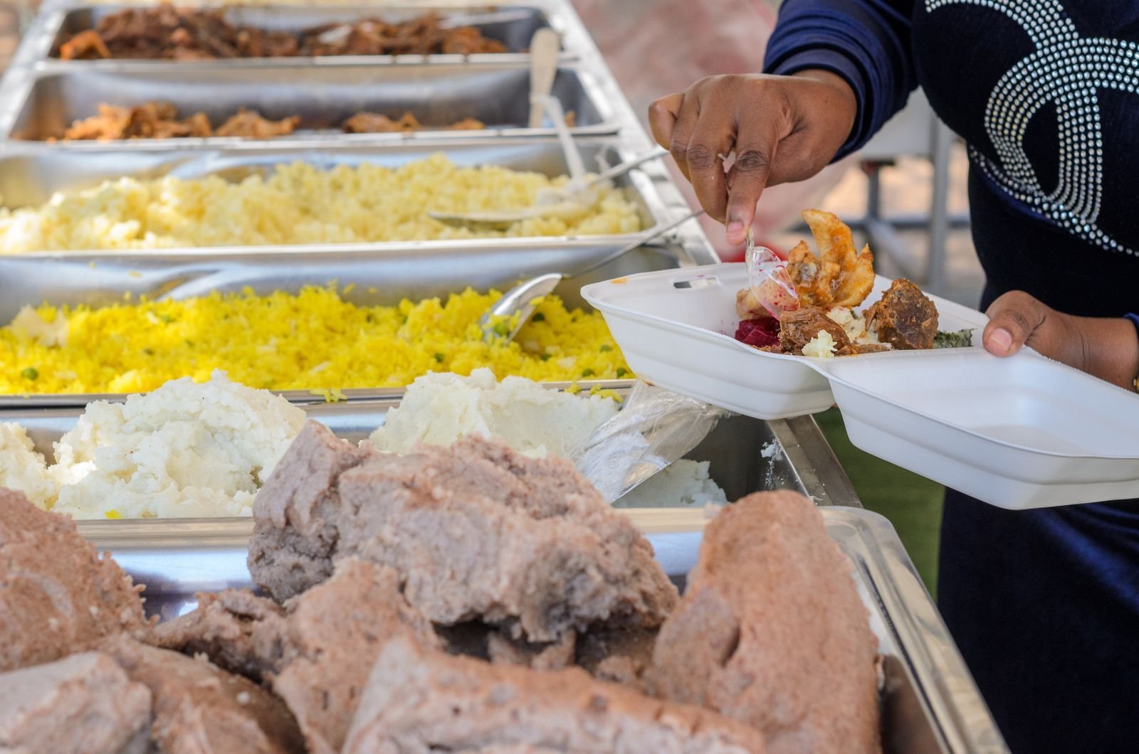 Pessoa se serve em balcão de restaurante self-service com cubas de arroz, carnes e feijão
