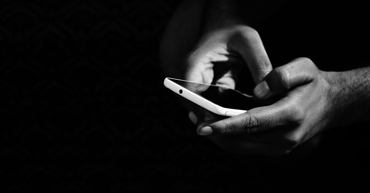 Mãos seguram um celular em foto em preto e branco, com fundo escuro