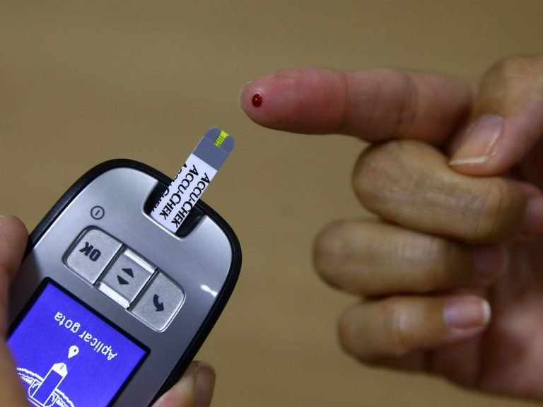 Aparelho medidor de glicose com tela em portugues ao lado de dedo com gota de sangue