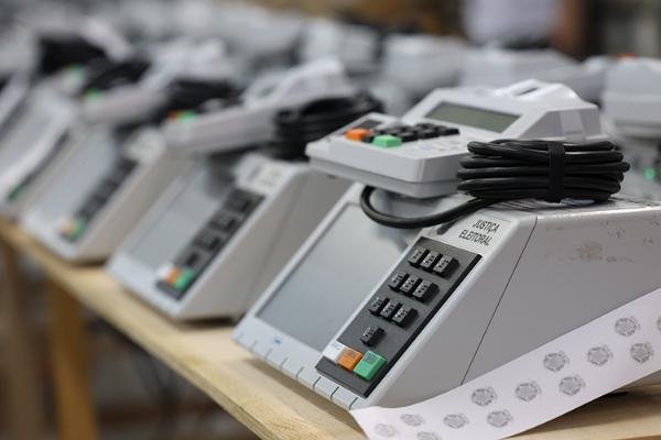 Urnas eletrônicas enfileiradas sobre bancada, com a inscrição Justiça Eleitoral