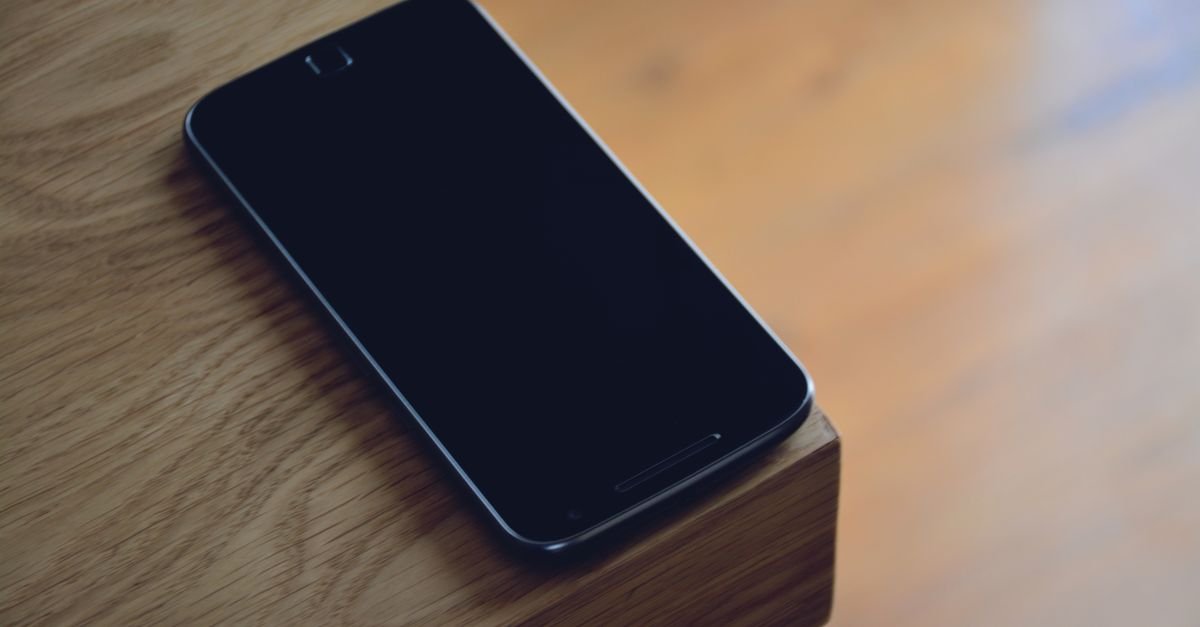 Celular com a tela apagada apoiado sobre a quina de uma mesa de madeira