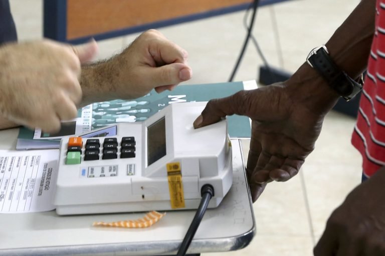Eleitor digita o voto em urna eletrônica durante votação, acompanhado por mesário