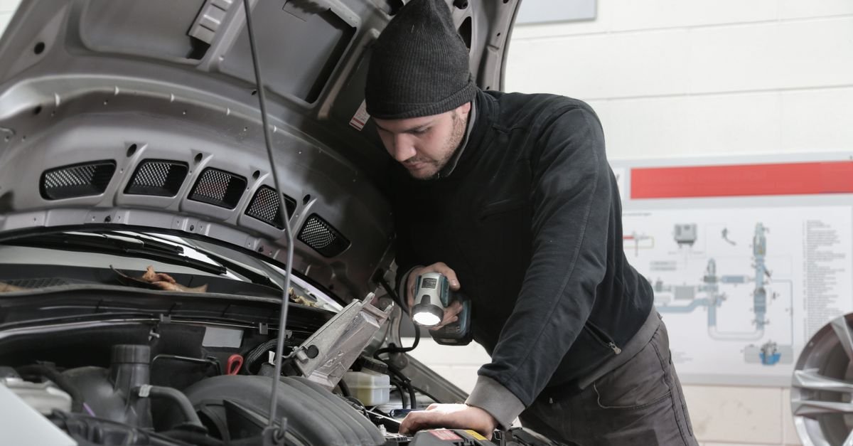 Mecânico com lanterna inspeciona o motor de um carro com o capô aberto em oficina